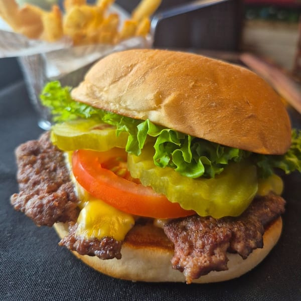 Cheeseburger with lettuce, tomato, and pickles on a bun, with fries in the background.