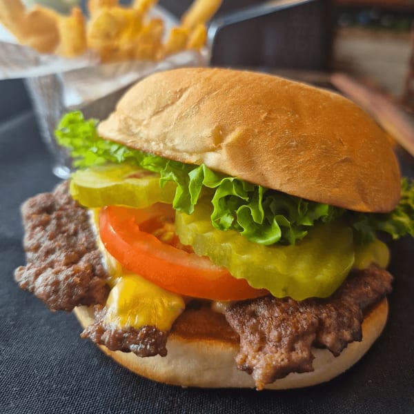 A Crazy Alan's Swamp Shack burger with lettuce, tomato, pickles, and cheese, with fries in the background.