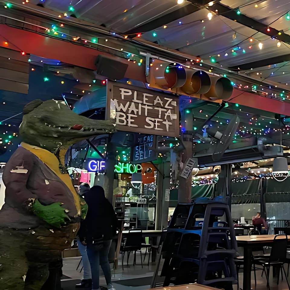 A large alligator statue in a suit stands next to a "Plez Wait to Be Set" sign at Crazy Alan's Swamp Shack.