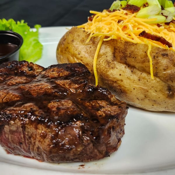 Grilled steak with a loaded baked potato, lettuce, and a side of sauce on a white plate.